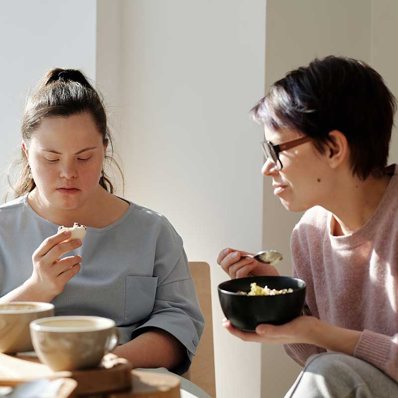 A young woman with Down's syndrome and her carer eating a meal in a sunny room and discussing helpful therapeutic supports from Mates Matter Counselling